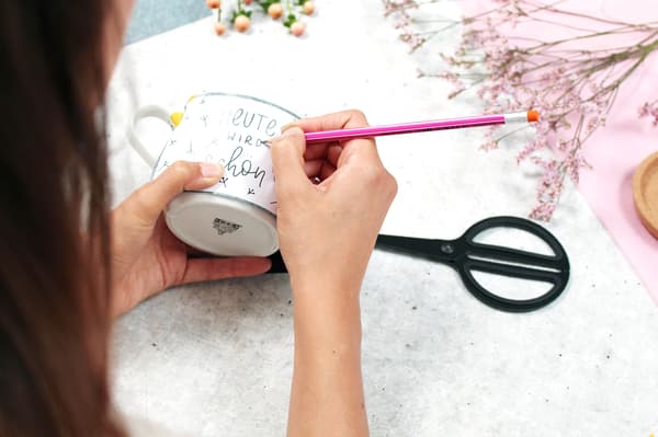 Jetzt nimmt die Frau einen Stift und zeichnet etwas auf die Tasse vor.