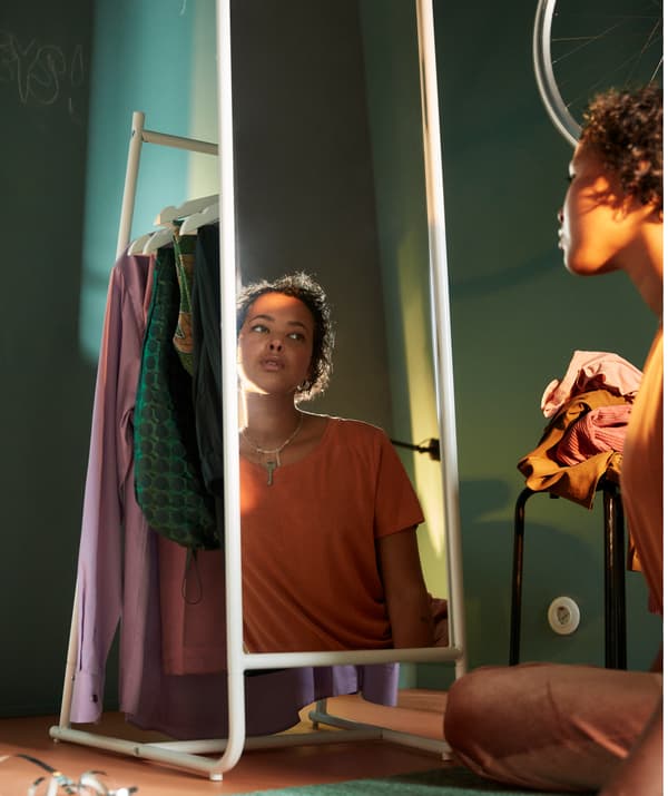 Jeune fille assise au sol se regardant dans un miroir. Une pile de vêtements est déposée sur un tabouret à côté d’elle.