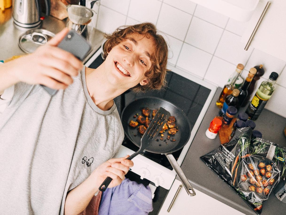 Joven sosteniendo un celular en su mano, tomándose una selfie donde apareerá su caray detrás un sartén con albóndigas IKEA dentro de él.