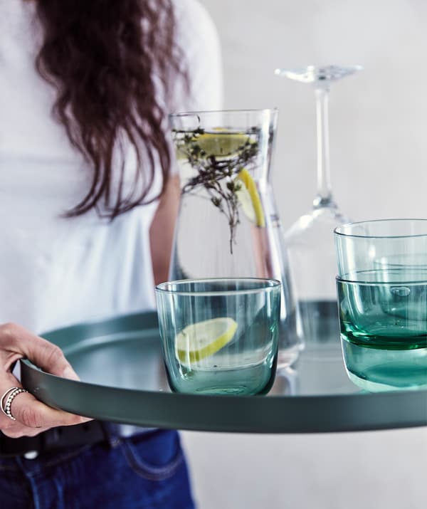 Julia hält GLADOM Tabletttisch in Dunkelgrün in Händen, darauf sind eine Karaffe mit Wasser und mehrere Gläser zu sehen.