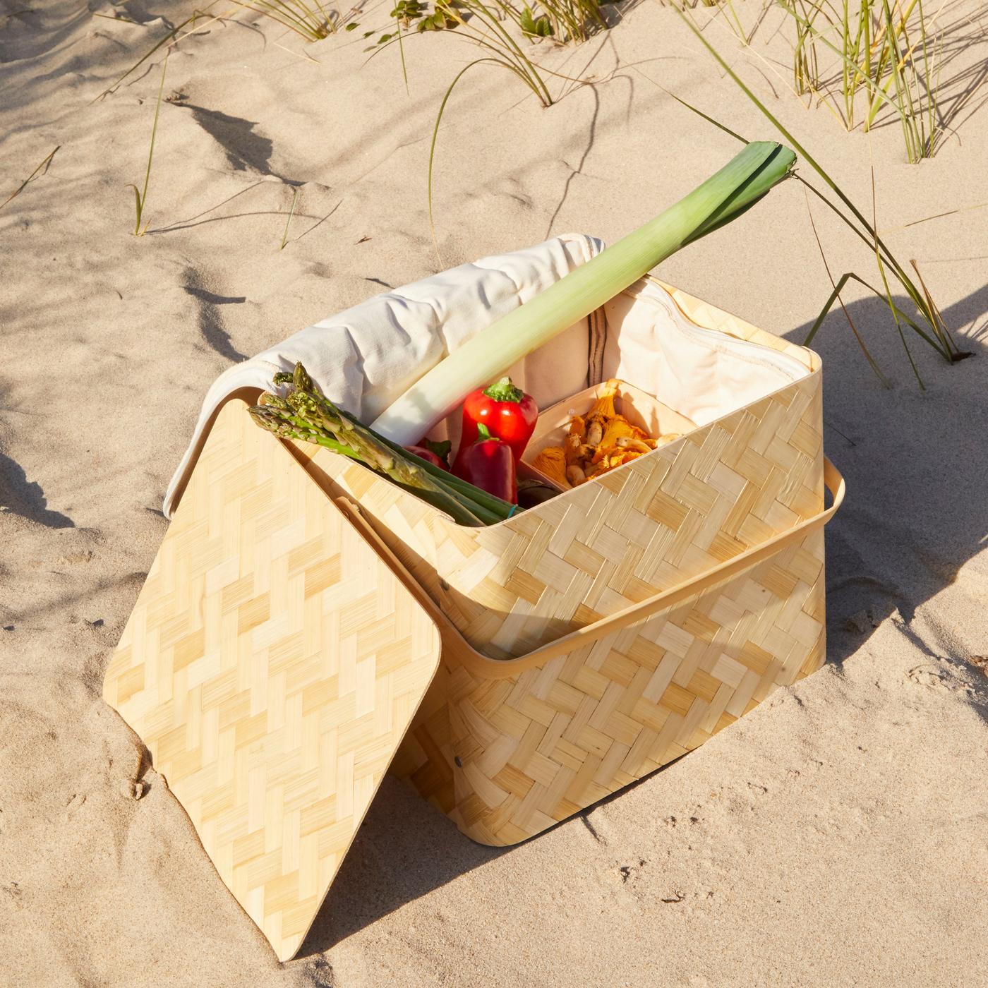 KÅSEBERGA cool basket made in woven bamboo and a cotton inside holding different vegetables, placed on a sandy beach.