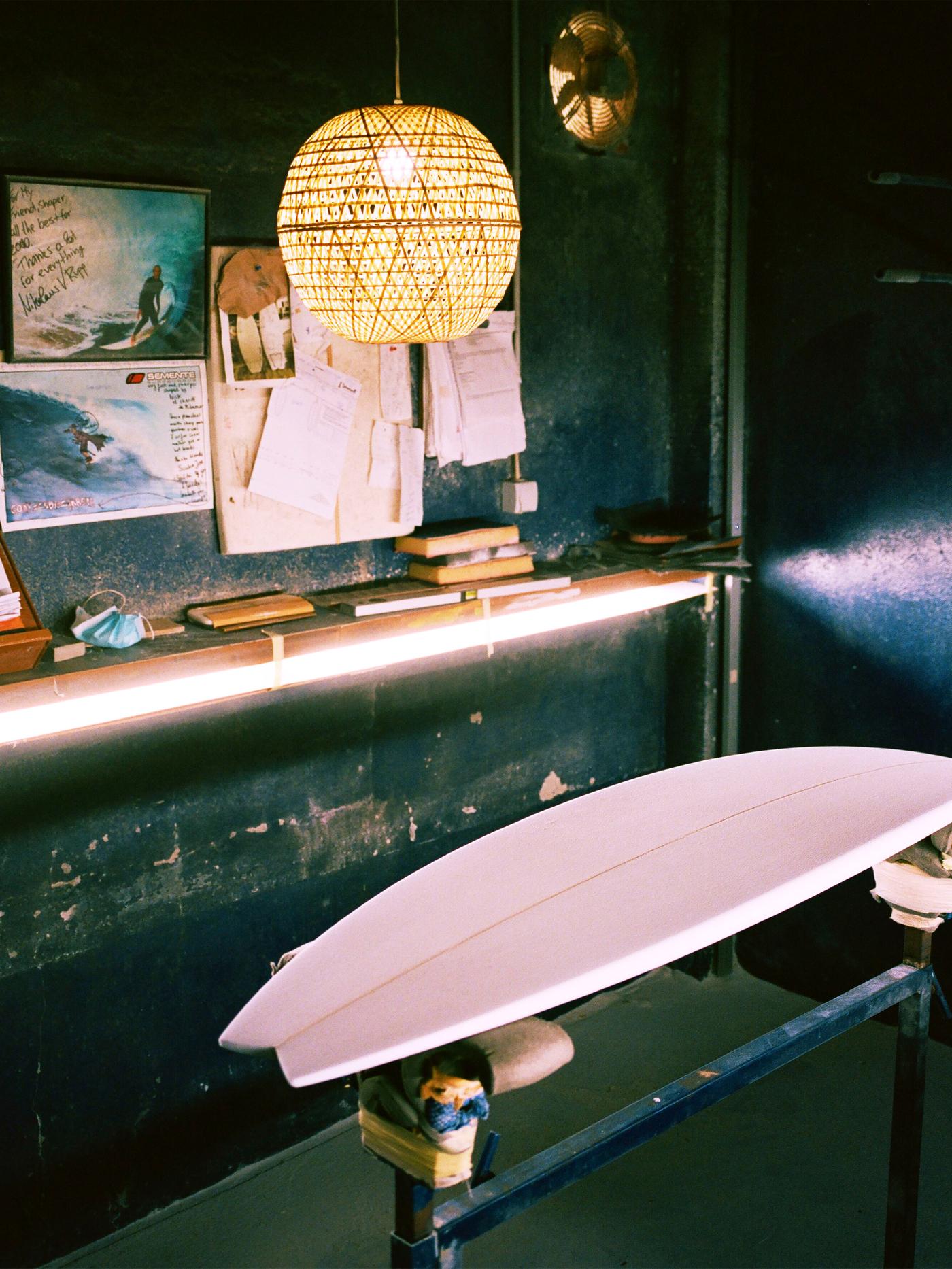 KÅSEBERGA lampshade made in woven bamboo hanging above a wooden surfboard placed in a dark-coloured artist’s workshop.