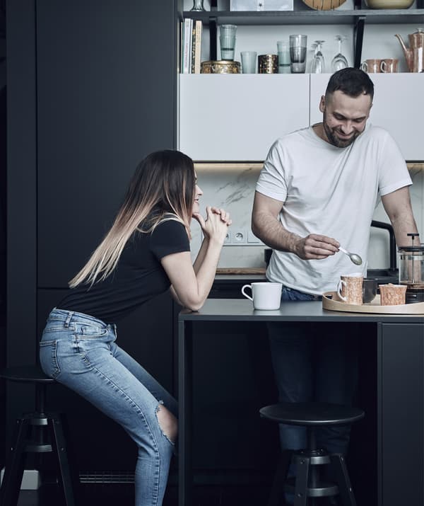 Katarzyna assise sur un tabouret de bar à l’îlot de sa cuisine noire, avec Mikolaj en train de préparer le café.