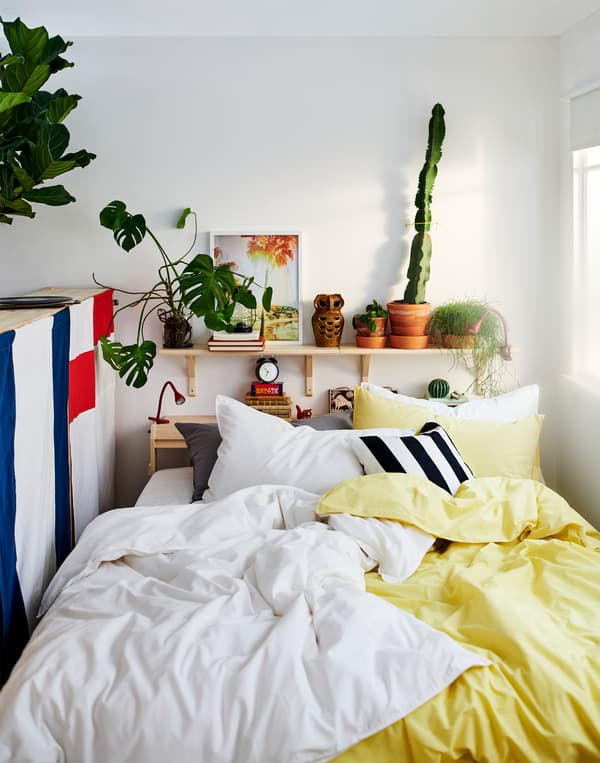 Katil dengan banyak bantal dan linen katil berwarna kuning, putih, hitam dan kelabu terletak di antara tingkap dan bahagian belakang unit storan.