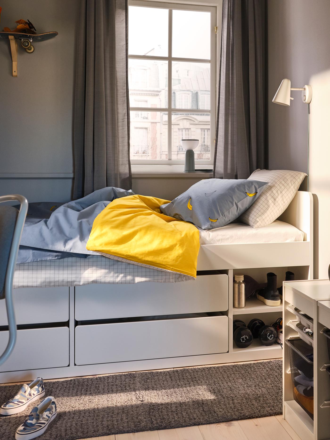 Katil dengan storan SLÄKT berwarna putih dengan linen katil VÄNKRETS terletak di bawah tingkap di dalam bilik tidur yang kecil.
