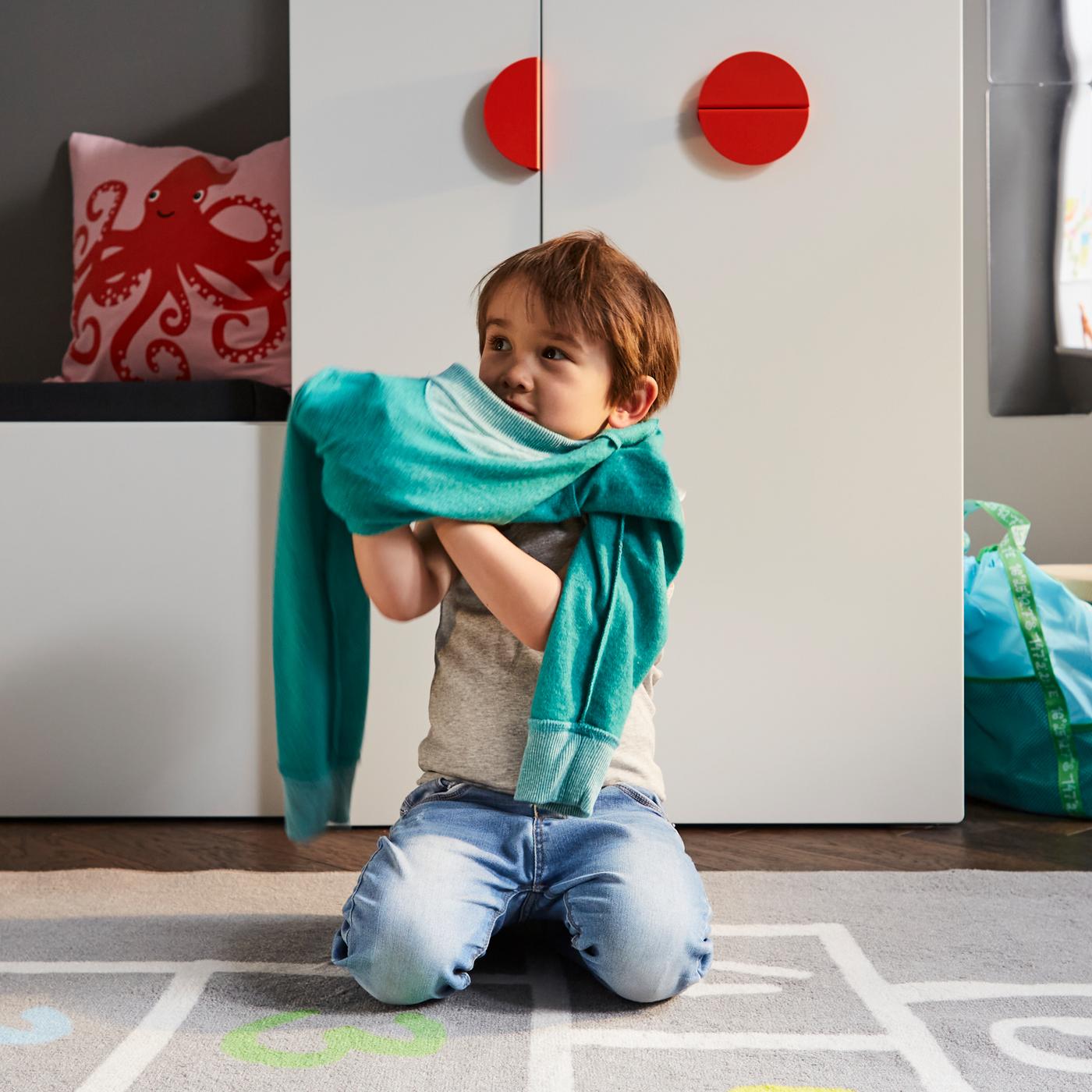 Kind kleedt zich aan op een grijs vloerkleed voor een witte bank met opbergruimte voor speelgoed en een witte kledingkast met uittrekbaar gedeelte.