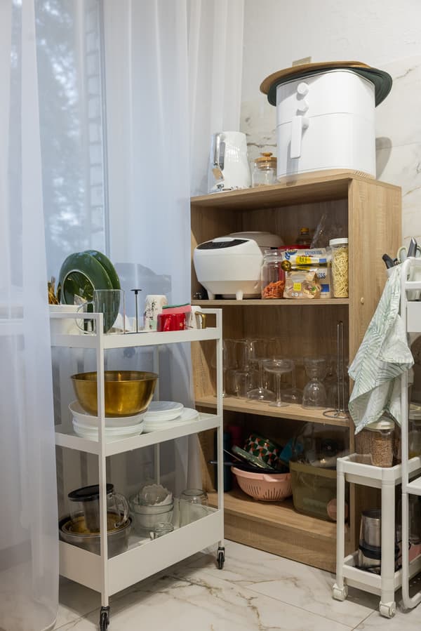 Kitchen corner with wooden and metal shelves holding dishes, food items and kitchen utensils; a window with light curtains is visible in the background.