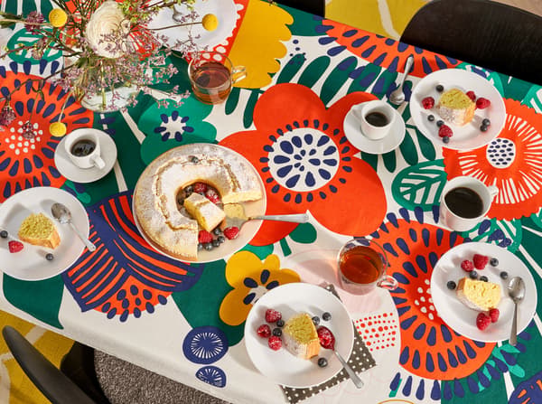 Kitchen with an overview of the kitchen table that has a flower-patterned tablecloth, plates, glasses, mugs and a cake on it.