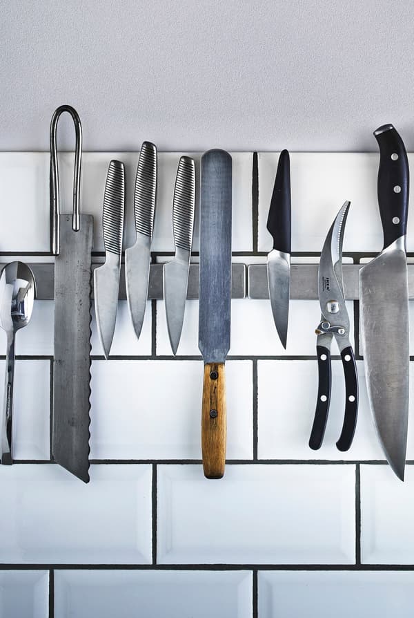 Knives and kitchen utensils stored on a magnetic rail mounted to the white tile wall in a kitchen.