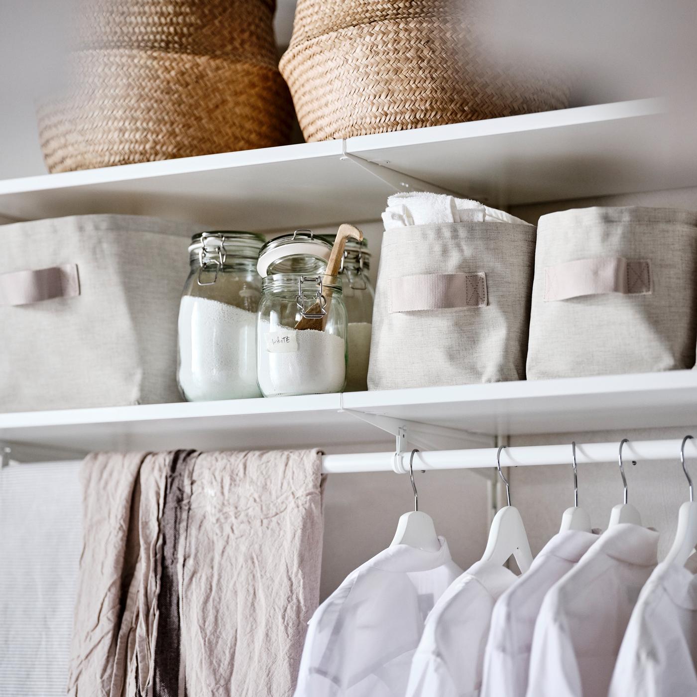 Kombinasi almari pakaian berwarna putih yang mempunyai bakul rumput laut, bakul storan tekstil berwarna kuning air dan pakaian pada penyangkut berwarna putih.