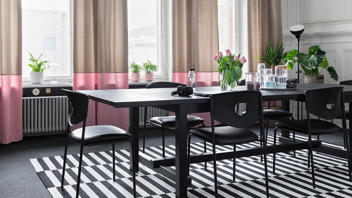 Konferensrum med stora fönster, beige och rosa gardiner, svart rektangulärt bord med svarta stolar på IKEA STOCKHOLM svartvit matta. På bordet finns en glasvas med rosa tulpaner och en bricka med en kanna med vatten, muggar och glas.