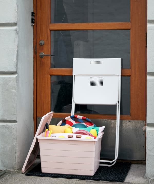 Kotak SOCKERBIT berwarna merah jambu yang penuh dengan keperluan untuk berkelah dan bermain terletak di pintu hadapan bangunan. Kerusi FEJAN yang boleh dilipat tersandar pada pintu.