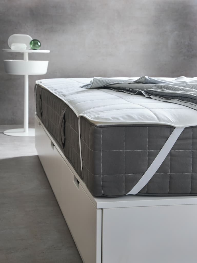 La esquina de un colchón gris sobre una cama de plataforma blanca con una pared gris en el fondo.