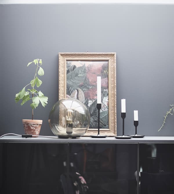 La lampe de table FADO apporte un peu de chaleur à la déco composée ici de bougies et d’une toile. L’abat-jour sphérique en verre gris clair diffuse la douce lumière de l’ampoule.