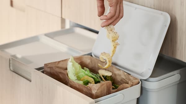 La mano de una persona metiendo una larga ristra de cáscaras de papas en un contenedor HÅLLBAR que contiene residuos orgánicos de cocina.