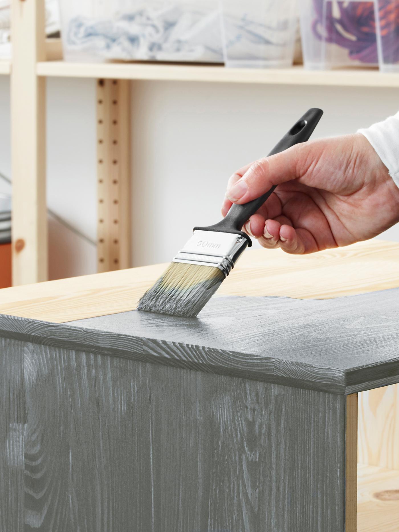 La mano de una persona sostiene un pincel y aplica pintura gris a la superficie superior de un mueble de pino sin tratar.