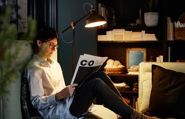 Lampadaire éclairant un journal lu par une femme assise sur le canapé.