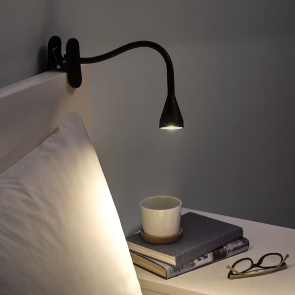 Lámpara de mesa NÄVLINGE negra sobre una cama y apuntando a dos libros y un par de anteojos de lectura.
