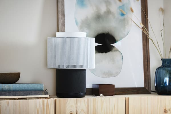 Lampe de table à haut-parleur intégré SYMFONISK déposée sur un chevet à proximité de nombreuses bougies. En arrière-plan, des rideaux blancs complètent le décor.
