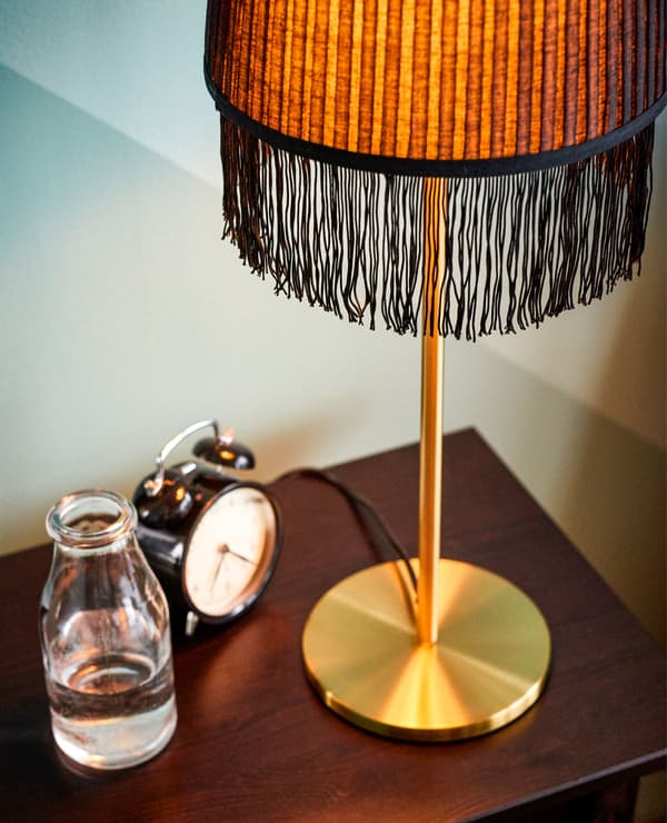 Lampe dorée à franges, réveil et verre d’eau, sur une table de chevet.