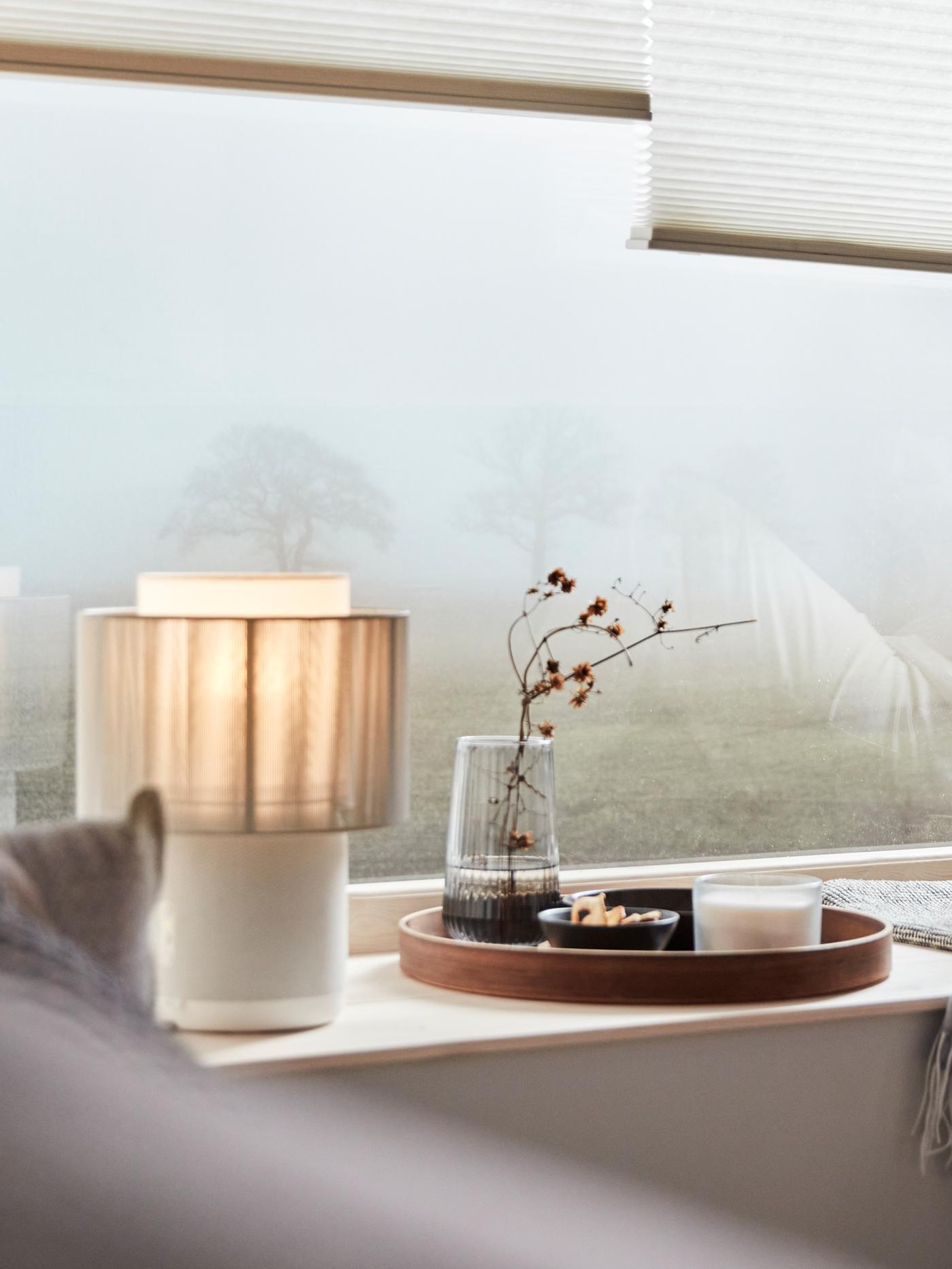 Lampe-enceinte SYMFONISK avec abat-jour textile posée sur un appui de fenêtre. Dehors, on distingue à peine un arbre dans la brume.
