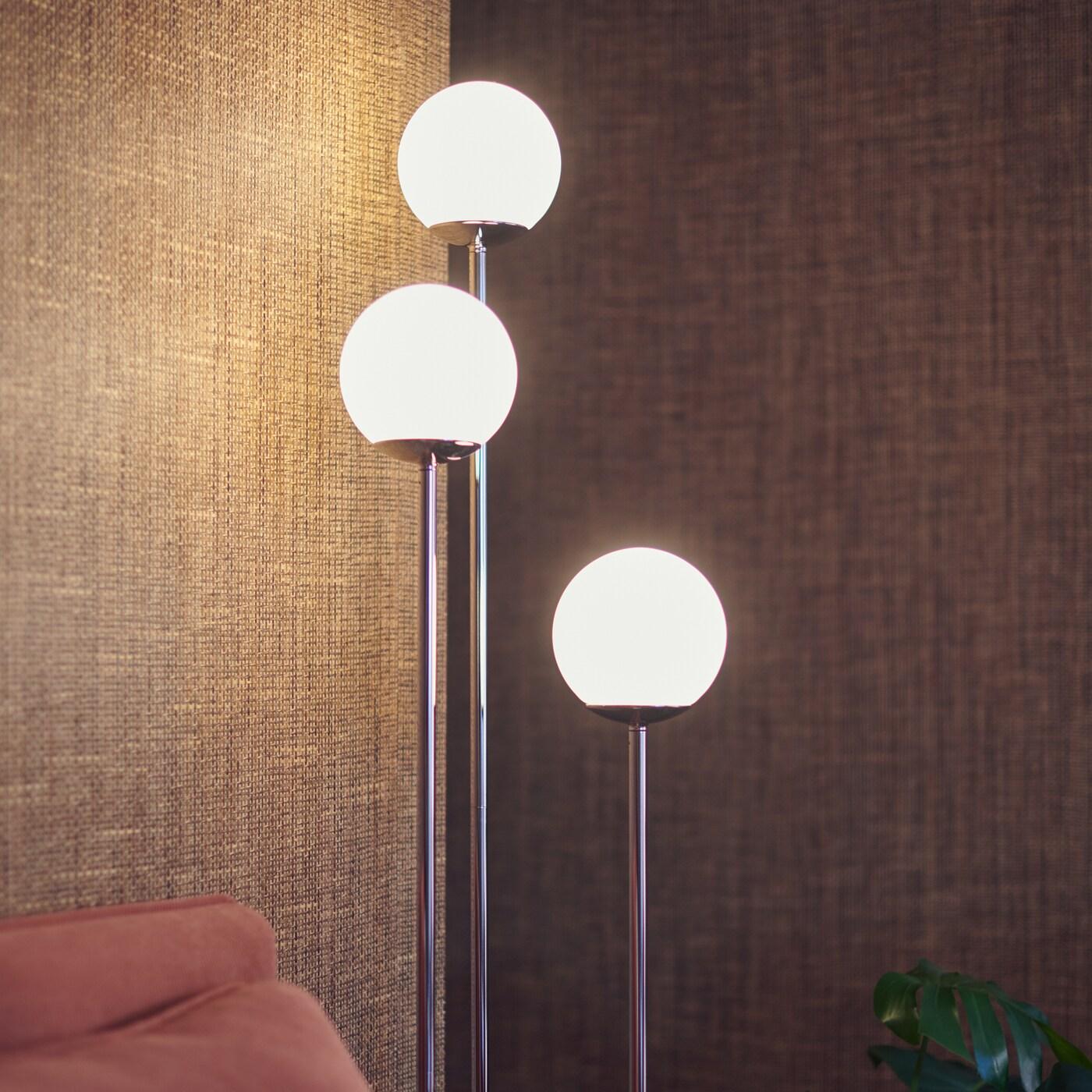 Lampu lantai SIMRISHAMN dengan tiga mentol memancarkan cahaya hangat pada dinding berwarna coklat semula jadi di penjuru ruang tamu.