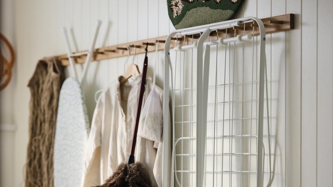 Laundry, cleaning and ironing accessories hanging from a row of HÖVOLM oak six-knob racks on a white, wood-panelled wall.