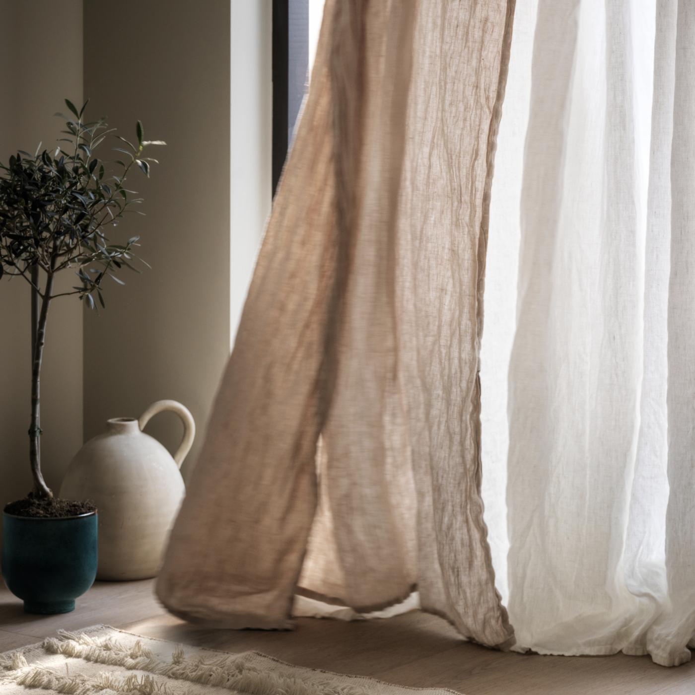 Le bas d’un rideau beige et d’un voilage SILVERLÖNN blanc effleure un plancher en bois et ondule devant une fenêtre.