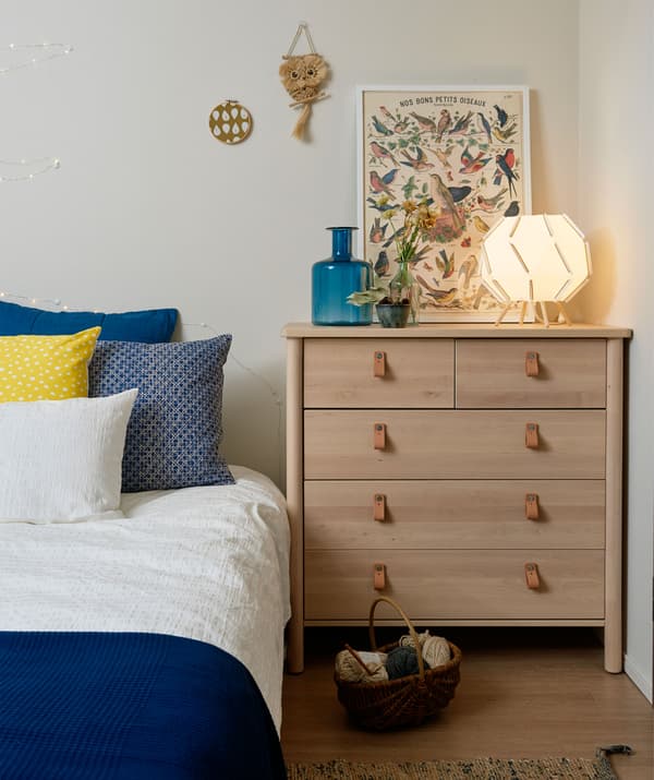 Le bord d’un lit avec linge de lit bleu et blanc à côté d’une commode dans une chambre blanche.