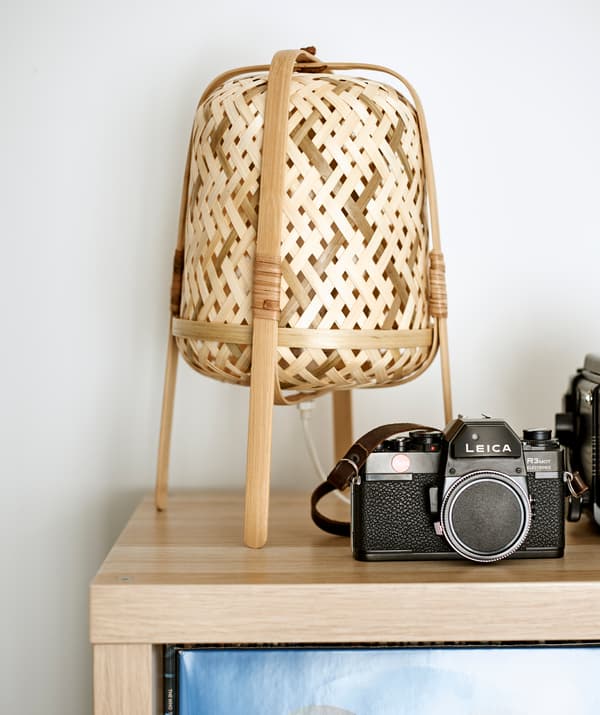 Le coin d’une étagère en bois clair au-dessus de laquelle sont posés un appareil photo vintage et une lampe de table en bambou tressé.