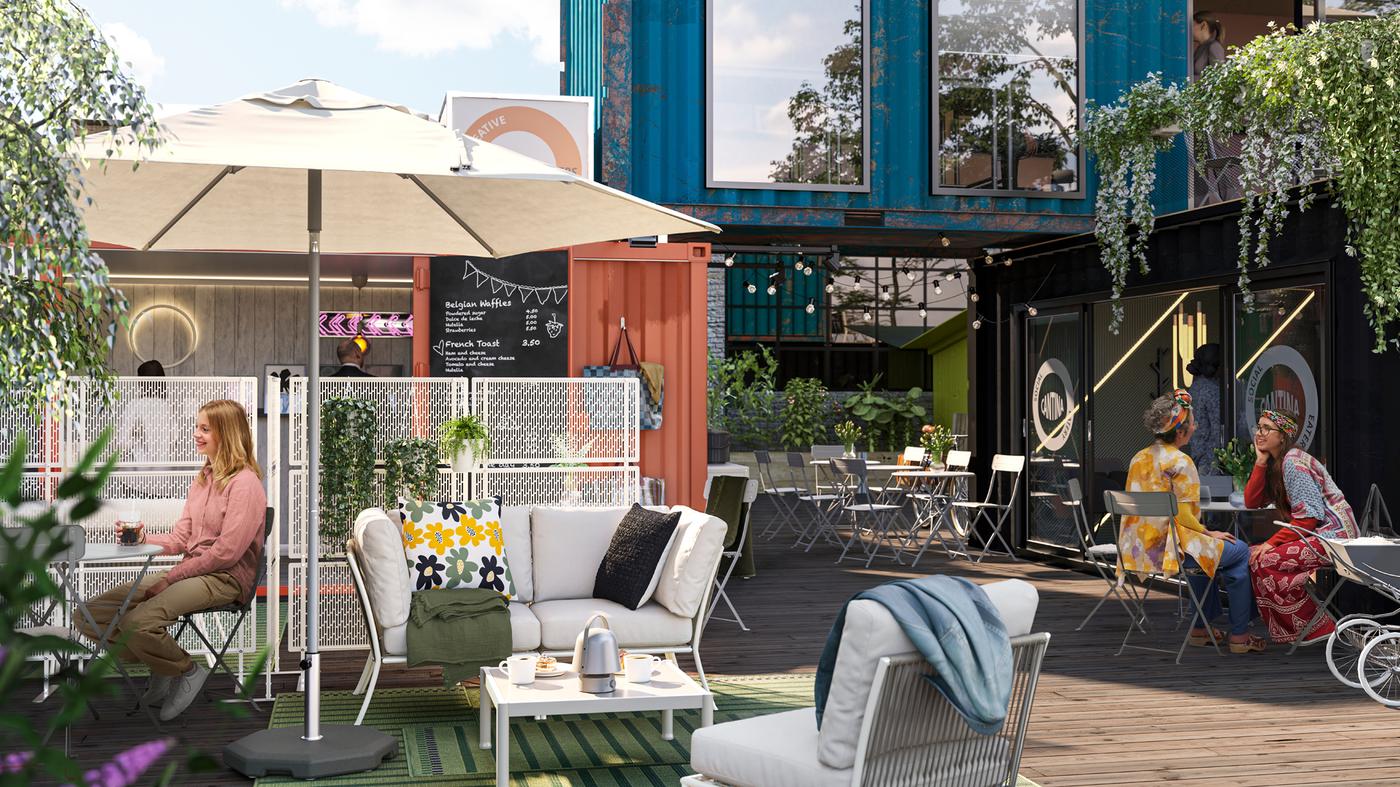 Le soleil brille sur un espace lounge extérieur dans un village de conteneurs. Des personnes discutent autour de tables SUNDSÖ grises.