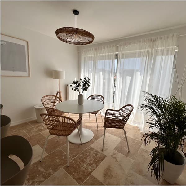 Lichte eetkamer met ronde witte tafel, rotan stoelen, transparante gordijnen, groene plant en hanglamp.