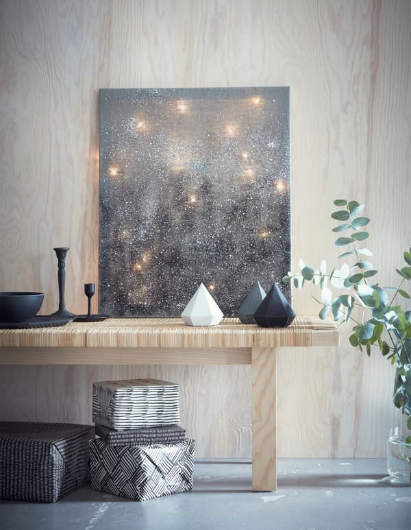 Light chains arranged behind a grey canvas, leaning on a light wood bench, with ornaments and gifts.