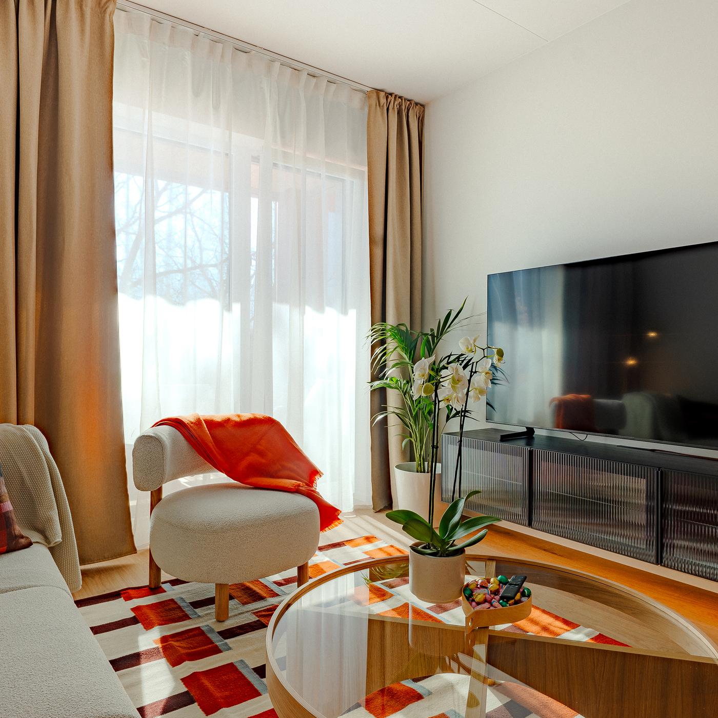 Light-filled living room with beige curtains, a sofa and armchair, a round glass coffee table, and a black BESTÅ TV bench with doors accented with terracotta textiles and plants.