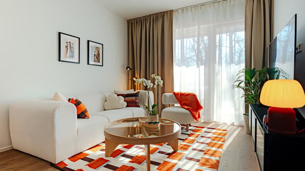 Light, Scandinavian‑inspired show apartment living room with a bright seating area, a neutral‑toned sofa and armchair, warm textile accents, and natural light from large windows.