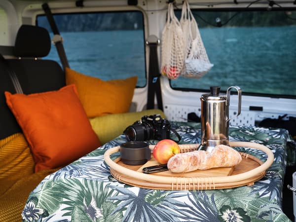 L'intérieur du van avec une vue de la table à manger avec une nappe à motif de jungle et une miche de pain, une pomme et une cafetière posées sur un plateau en bois. 