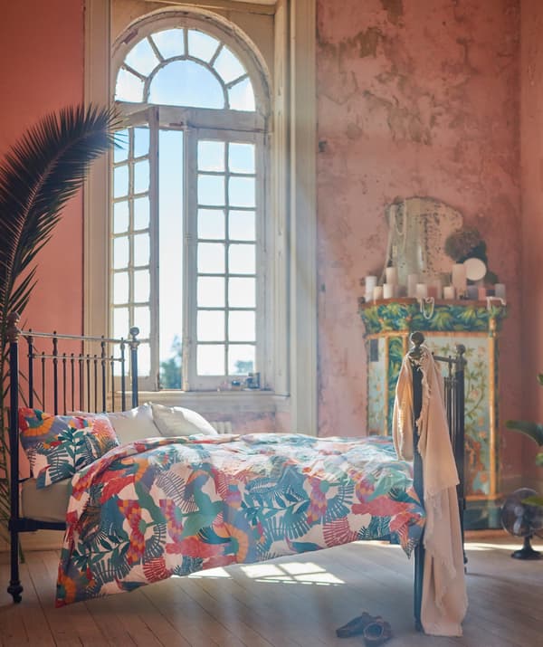 L’intérieur d’une chambre éclairée par une grande fenêtre en arc. Un lit à structure métallique extra-plate est fait avec du linge de lit SKOGSFIBBLA.