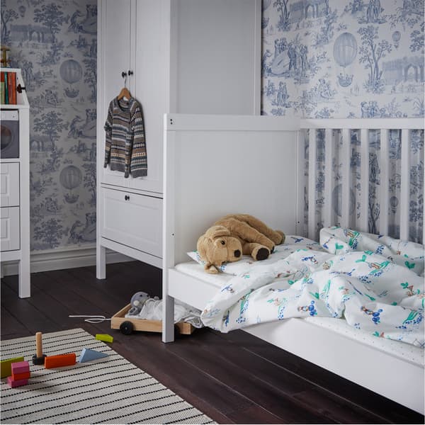 Lit bébé blanc avec du linge de lit blanc/à motif et un chien en peluche, des jouets en bois au sol, une garde-robe en arrière-plan.