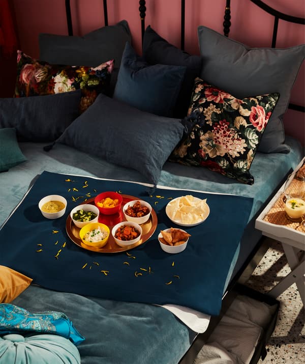 Lit recouvert d'une couverture et beaucoup de coussins sur lequel est servi un repas posé au milieu d'un morceau de tissu qui sert de nappe.