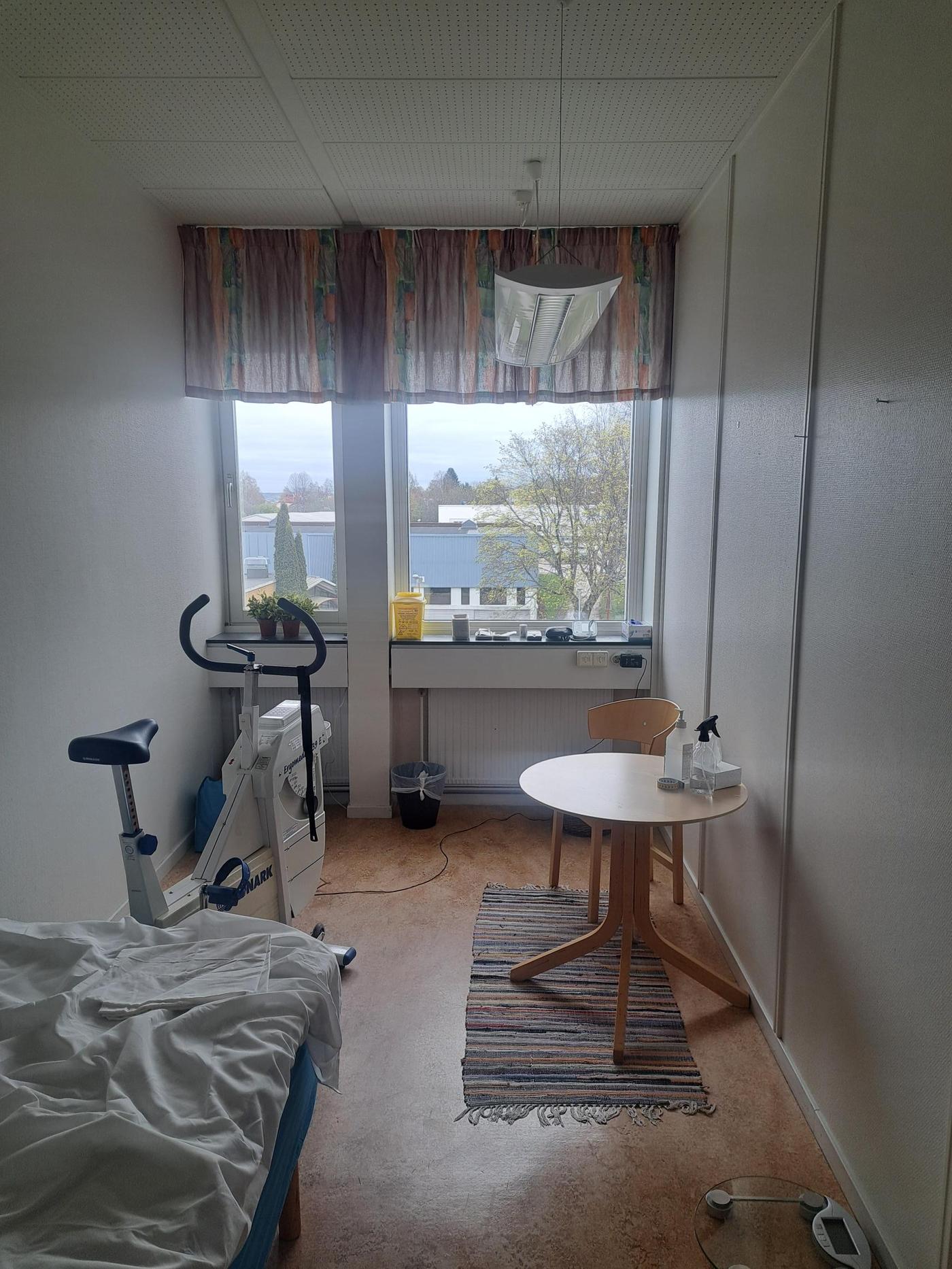 Litet, välstädat rum med säng, motionscykel och ett runt bord med två stolar. Färgglada gardiner ramar in två stora fönster som släpper in dagsljus och visar utsikt över träd och byggnader