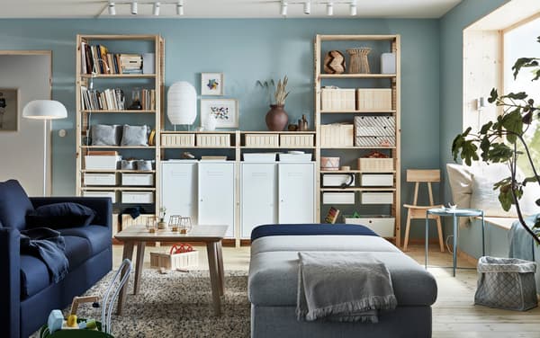 Living donde hay un sofá azúl oscuro VIMLE a la izquierda, una mesa de centro de madera y módulos de sofá grises y azul para sentarse a la derecha. Un estante grande atrás de madera lleno de libros y cajas organizadoras.