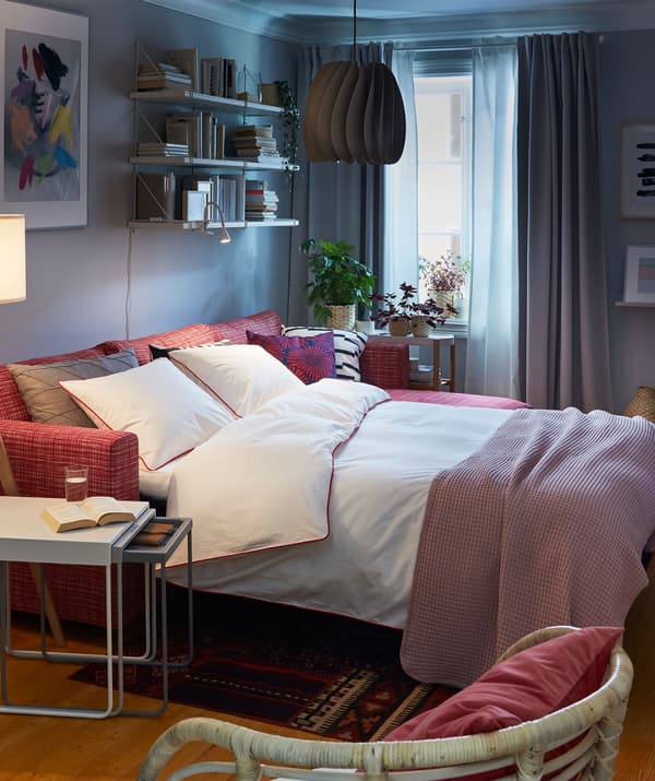 Living room interior with sofa bed in double-bed mode. Around it: a nest of tables, seating, wall-mounted bookcase with books.