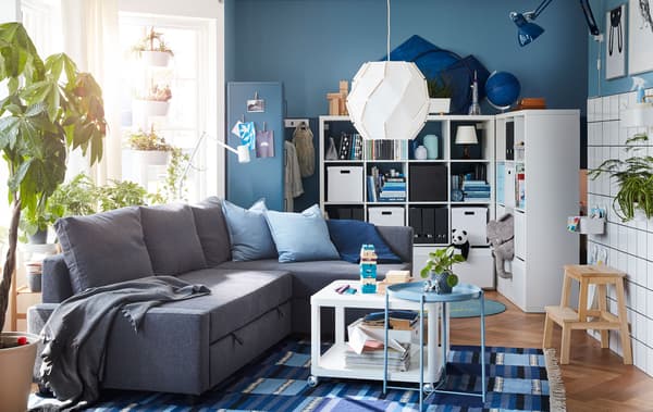 Living room with a corner sofa at centre, surrounded by large, filled shelving units, big windows and many plants.