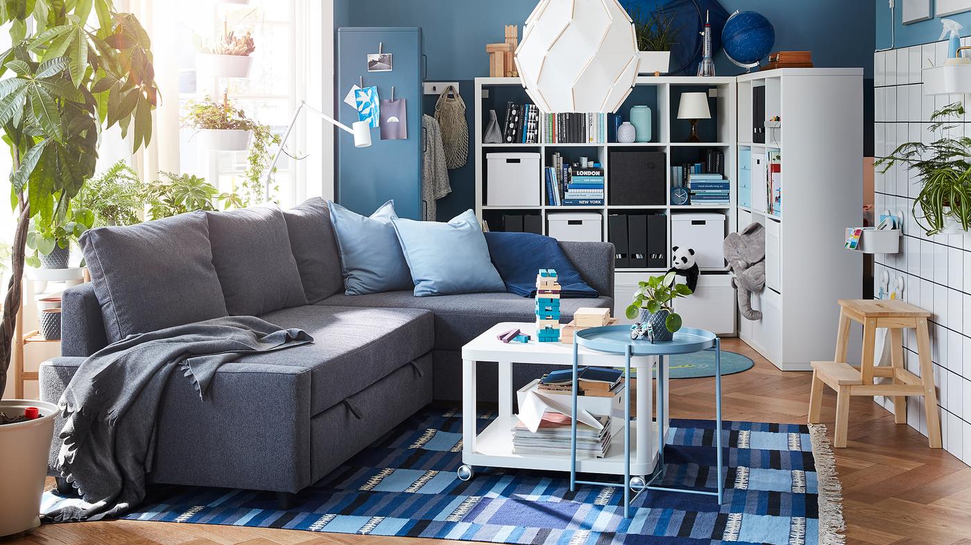 Living room with a dark grey corner sofa-bed, a blue-patterned rug and a freestanding shelving unit with space behind it.