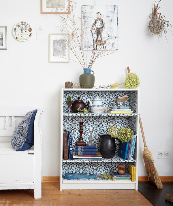 Livres et décorations sur une petite bibliothèque avec intérieur à motif floral, dans une pièce aux murs blancs et au plancher en bois.