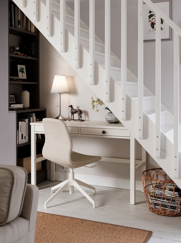 Located under a staircase is a workspace consisting of a white HEMNES desk, a LÅNGFJÄLL chair, a lamp and a tall bookcase.