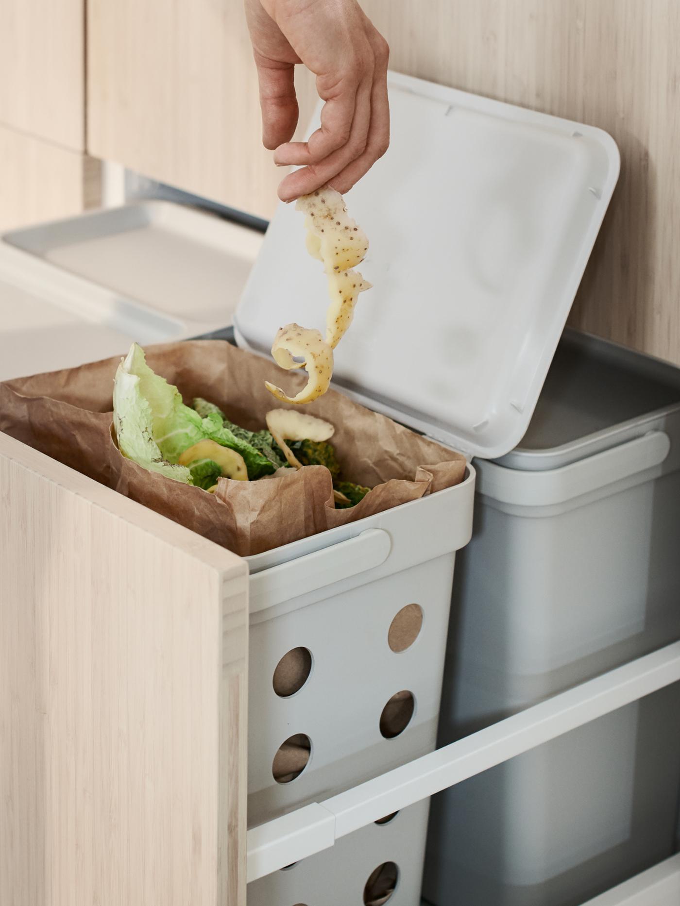 Main tenant une longue pelure de pomme de terre au-dessus d’une poubelle HÅLLBAR contenant des déchets de cuisine.