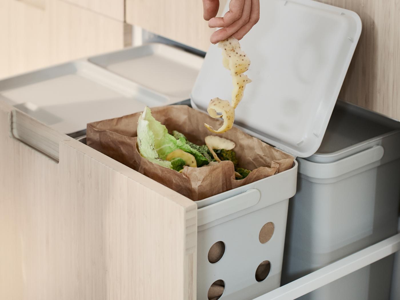 Main tenant une longue pelure de pomme de terre au-dessus d’une poubelle HÅLLBAR contenant des déchets de cuisine.
