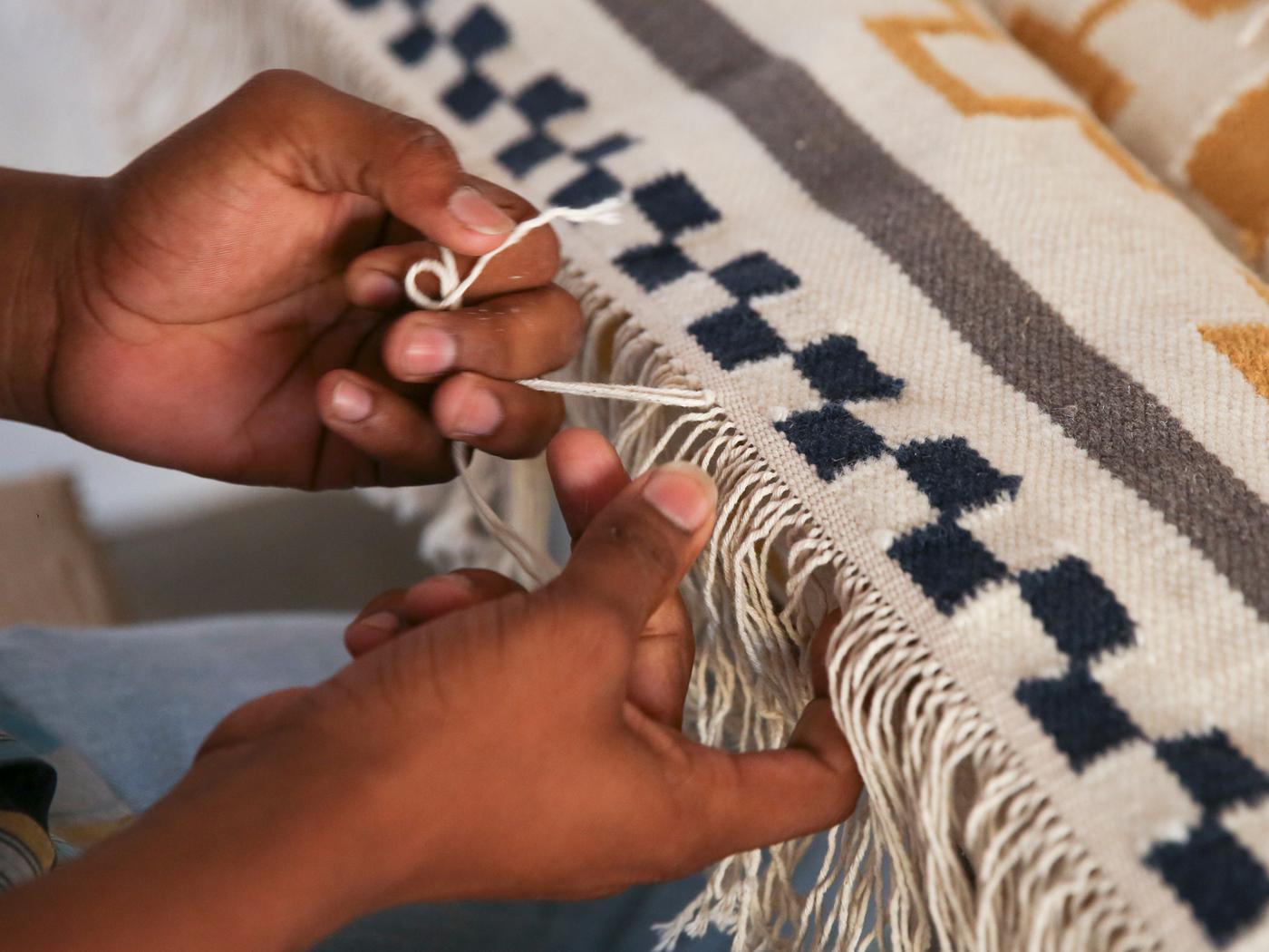 Mains tissant la frange d’un tapis dans des tons de blanc, bleu, gris et moutarde.