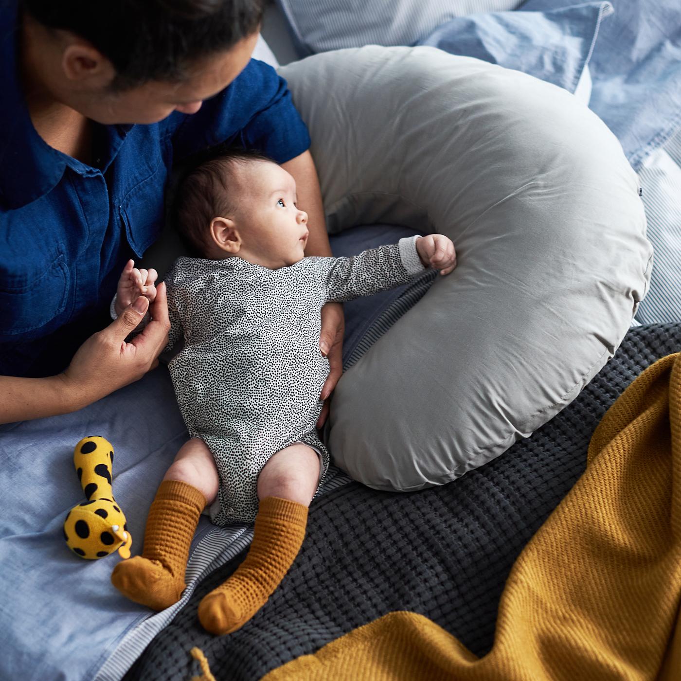 Majka u krilu drži bebu u bodiju prekrivenom tačkicama, a pored bebe je žuta/crna KLAPPA zvečka.